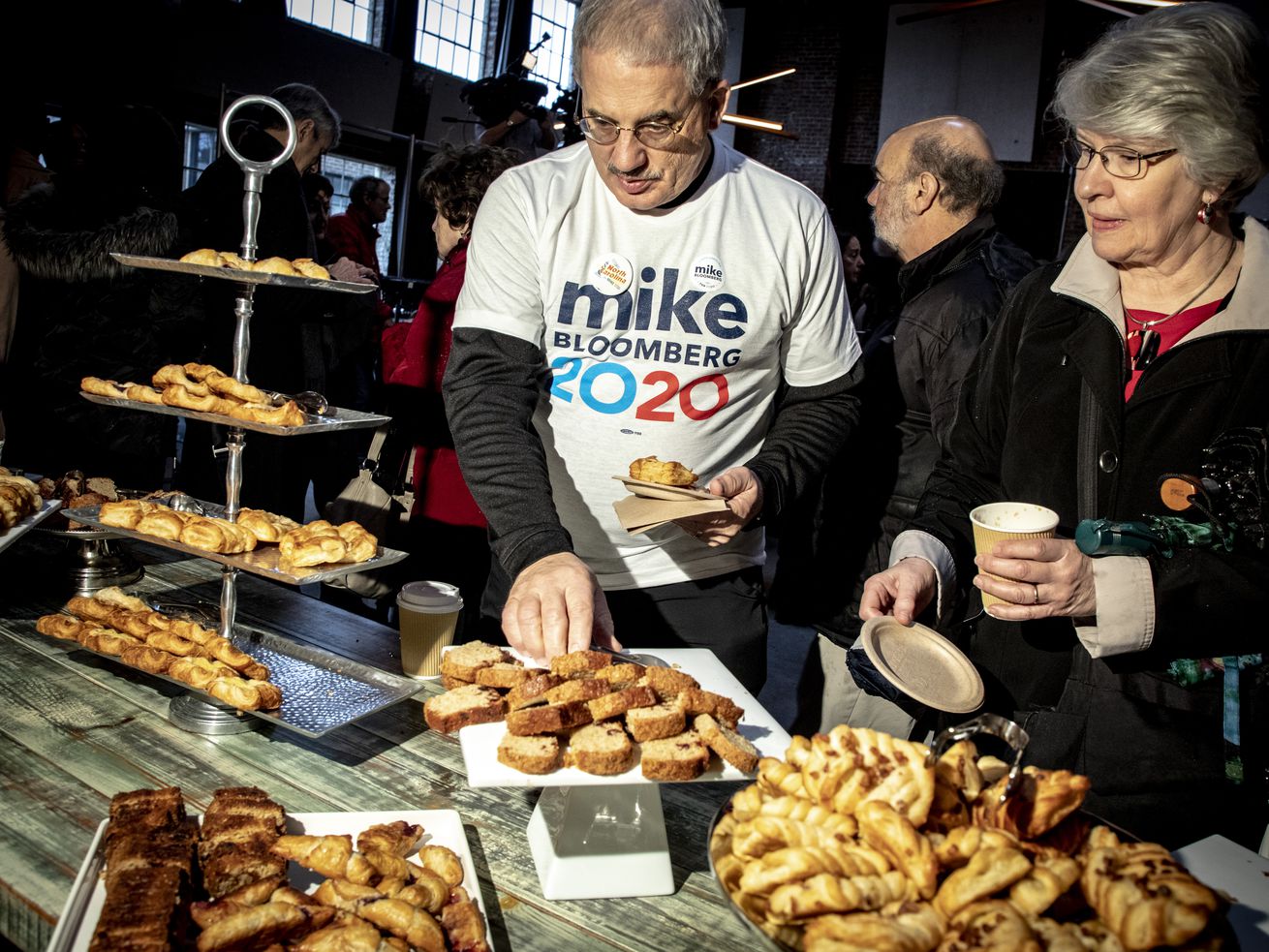 Man and woman look like a table filled with pastries; the man wears a shirt that reads “Mike Bloomberg 2020.”