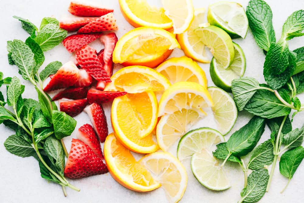 Photo filled with a mint, sliced strawberries, quartered oranges, lemons and limes with another side of mint on a table. 